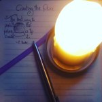A birds-eye view of a lit, large cream candle, lighting a stainless steel pen and a journal page with a quote "the best way to predict the future is to create it." and a purple ribbon bookmark.