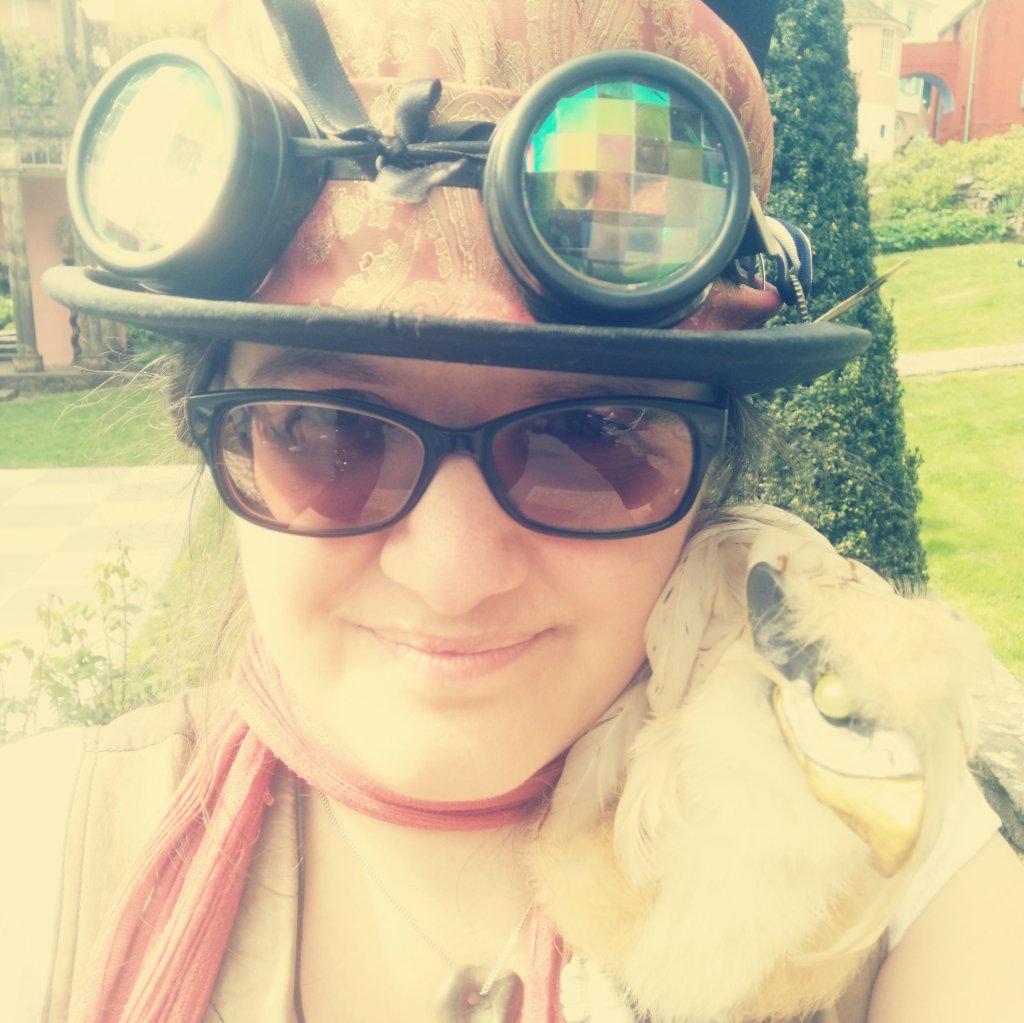 Halo (a white femme in tinted glasses and a top hat with kaleidoscope lensed goggles) at a Steampunk event, grinning at the camera with a small cream coloured gryphon (half lion, half eagle) on their shoulder!